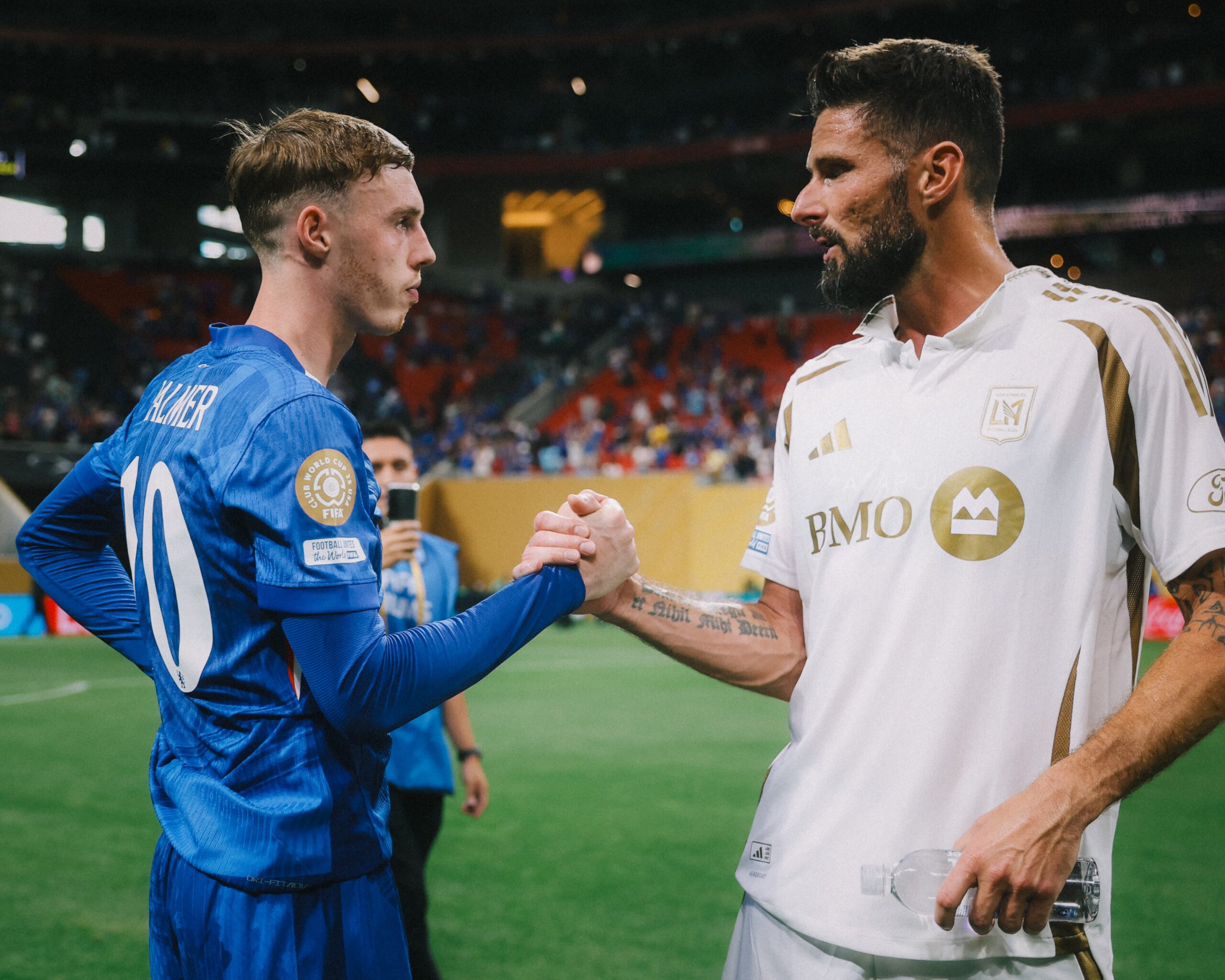 Cole Palmer and Olivier Giroud shaking hands on the Mercedes-Benz Stadium pitch, both in their respective kits, with the crowd in the background before Chelsea’s Club World Cup 2025 match against LAFC.
